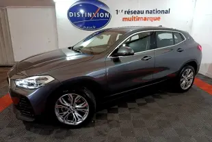 BMW X2 sDrive18i gris foncé vue de profil côté gauche, avec jantes alliage et phares allumés dans un showroom.