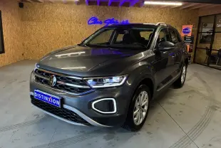 Volkswagen T-Roc gris 3/4 avant droit, phares allumés, calandre chromée et jantes alliage visibles en intérieur showroom.