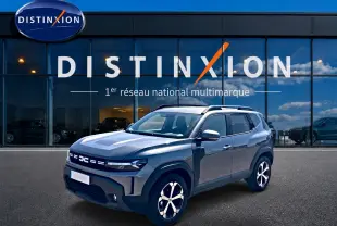 Dacia Duster gris schiste en 3/4 avant droit, avec jantes alu et barres de toit, devant une vitrine Distinxion.