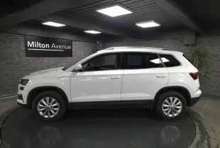 Profil côté gauche du SUV blanc SKODA KAROQ 2024 en intérieur avec jantes alliage et barres de toit chromées.
