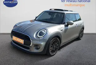 Vue 3/4 avant d'une MINI beige avec jantes noires et bandes décoratives sur le capot, stationnée sur sol gris.
