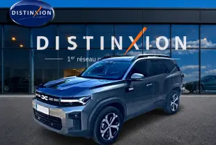 SUV Dacia Bigster gris schiste en 3/4 avant droit, avec jantes alu et barres de toit visibles devant un showroom Distinxion.