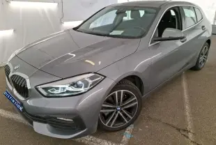 BMW Série 1 gris vue 3/4 avant droit, phares allumés et jantes alliage distinctives, stationnée en intérieur.