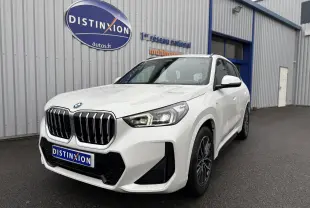 BMW X1 18D blanc minéral en vue 3/4 avant droit, avec calandre imposante et phares LED allumés.