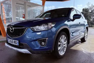 Vue 3/4 avant droit d'un Mazda CX-5 bleu 2013 avec calandre noire et jantes argentées, sous un abri extérieur.