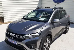 Dacia Sandero Stepway gris schiste vue 3/4 avant droit, avec barres de toit et stickers publicitaires visibles.