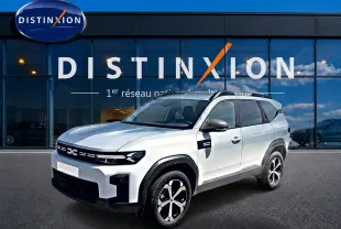 Vue 3/4 avant droit d'un Dacia Bigster blanc avec barres de toit et jantes alliage devant un bâtiment vitré Distinxion.