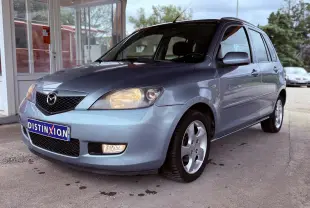 Vue 3/4 avant droit d'une Mazda2 gris clair avec phares allumés, garée devant un bâtiment vitré.