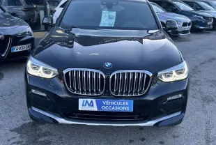 BMW X4 X Drive gris foncé vue de face avec calandre imposante et phares allumés sur parking extérieur