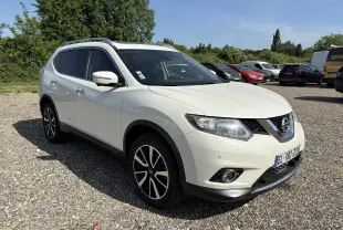 Nissan X-Trail 2016 blanc en 3/4 avant droit, avec jantes alliage et barres de toit visibles sur un sol gravillonné.