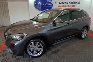 BMW X1 sDrive18i gris foncé vue 3/4 avant droit, feux allumés, roues alliage et rails de toit noirs visibles