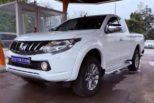 Mitsubishi L200 Club Cab blanc vu en 3/4 avant droit avec calandre noire et phares allumés sous abri.