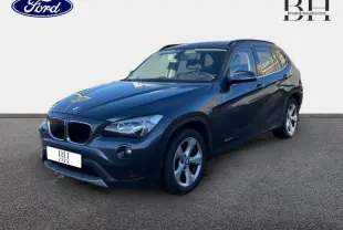 BMW X1 sDrive20d 2013 Mineralgrau en 3/4 avant droit avec jantes alu 17 pouces et barres de toit noires mat.