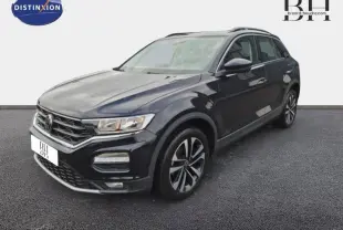 Volkswagen T-Roc gris en 3/4 avant droit, avec jantes alliage et barres de toit visibles.