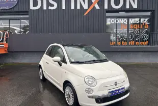Fiat 500 blanc vue 3/4 avant droit sous la pluie, toit noir et jantes alliage 15 pouces visibles.